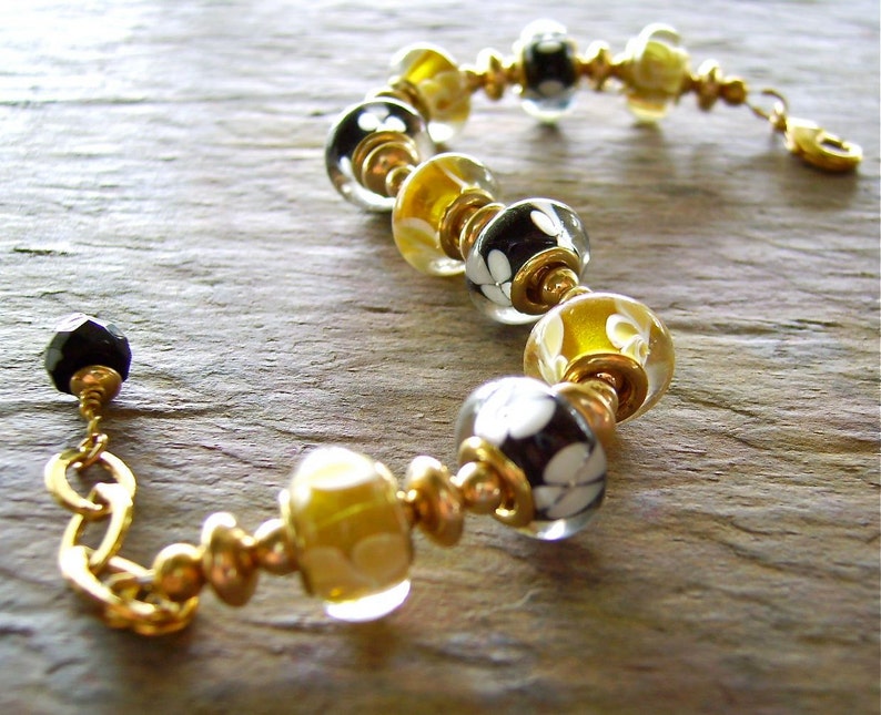 May include: A bracelet featuring alternating glass beads in black and yellow, each decorated with white floral patterns. The beads are separated by small gold-toned metal spacers and rings, finished with a gold-toned lobster clasp and a black bead.