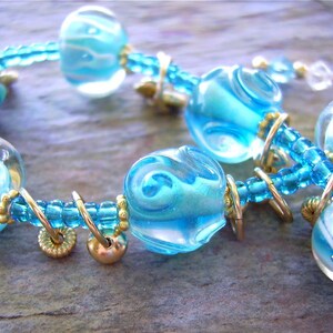 K&ouml;nnte beinhalten: Ein Armband mit durchscheinenden blau-wei&szlig;en Wirbelglasperlen, durchsetzt mit kleineren hellblauen Saatperlen und goldfarbenen Metallringen. Das Design umfasst eine gr&ouml;&szlig;ere lavendelfarbene und blaue Glasperle sowie goldfarbene Zwischenelemente.