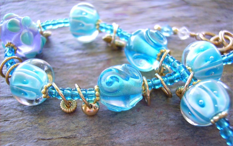 K&ouml;nnte beinhalten: Ein Armband mit durchscheinenden blau-wei&szlig;en Wirbelglasperlen, durchsetzt mit kleineren hellblauen Saatperlen und goldfarbenen Metallringen. Das Design umfasst eine gr&ouml;&szlig;ere lavendelfarbene und blaue Glasperle sowie goldfarbene Zwischenelemente.
