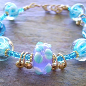 K&ouml;nnte beinhalten: Ein Armband mit durchscheinenden hellblauen und wei&szlig;en Wirbelglasperlen, kleinen leuchtend blauen Saatperlen und goldfarbenen Metallringen. Eine zentrale lavendelfarbene Glasperle mit hellblauen und wei&szlig;en Blumenmustern wird von goldfarbenen Metallabstandshaltern flankiert.