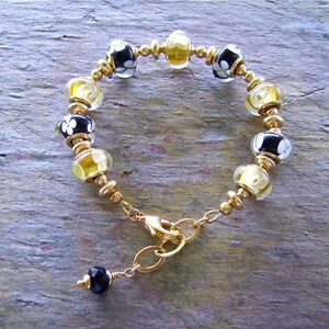 May include: A bracelet featuring alternating black glass beads with white floral patterns and translucent yellow glass beads, separated by small gold-toned metal spacers. The piece includes a gold-toned lobster clasp and a small black bead charm.
