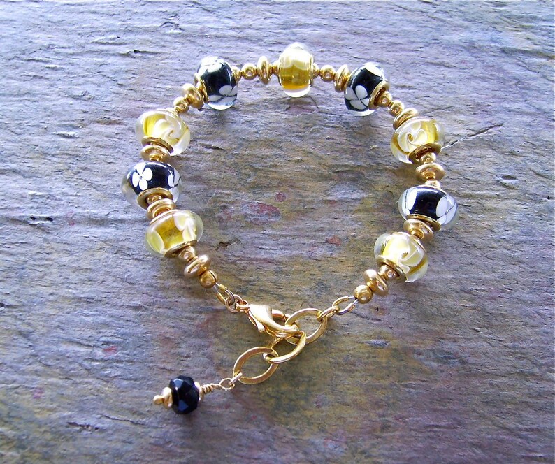 May include: A bracelet featuring alternating black glass beads with white floral patterns and translucent yellow glass beads, separated by small gold-toned metal spacers. The piece includes a gold-toned lobster clasp and a small black bead charm.