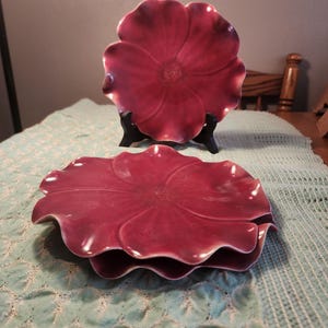 May include: Two burgundy-colored, flower-shaped plates. One plate is displayed on a black stand, while the other is stacked below. The plates have a glossy finish and a detailed floral design. They are placed on a light blue patterned tablecloth.