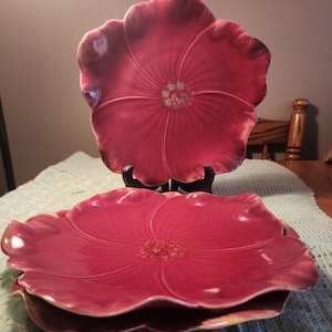 May include: A stack of three red ceramic plates shaped like flowers. The plates have a glossy finish and a detailed floral design. One plate is displayed on a black stand. The plates are a vibrant shade of red.
