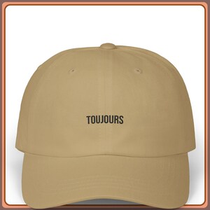 Può includere: Un cappellino da baseball beige con visiera curva. La parola "TOUJOURS" è ricamata in nero al centro. Il cappellino ha una corona strutturata e un cinturino regolabile.