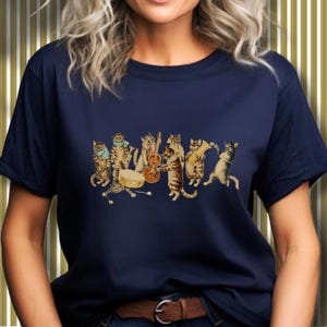 Może przedstawiać: Granatowa koszulka z zabawną ilustracją zespołu kotów. Zespół składa się z kilku kotów grających na różnych instrumentach, w tym na gitarze, perkusji i puzonie. Koty są przedstawione w stylu vintage.