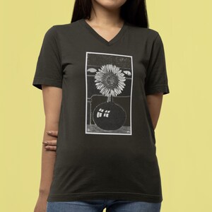 May include: Dark green V-neck t-shirt featuring a black and white illustration of a sunflower in a vase. The graphic is rectangular and centered on the shirt. The shirt has short sleeves and a relaxed fit.