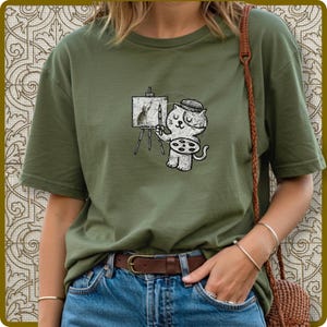 May include: Olive green t-shirt featuring a cartoon cat artist. The cat, wearing a beret, holds a paintbrush and palette, painting on a small easel. The artwork on the easel depicts a rabbit. The shirt is paired with blue jeans and a brown belt.