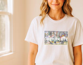 Puede incluir: Camiseta blanca con un diseño rectangular con el texto "COO YORK CITY". El diseño muestra un paisaje urbano de dibujos animados con palomas. La camiseta está hecha de un material suave.