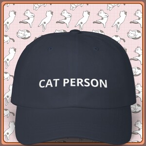May include: A navy blue baseball cap with the embroidered text "CAT PERSON" in white. The cap has a curved brim and a structured crown. The background features a repeating pattern of cartoon cats.