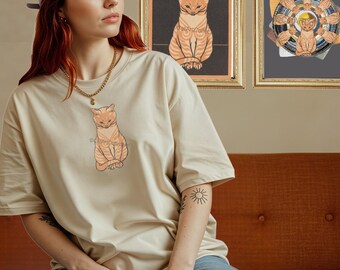 May include: Beige t-shirt featuring a seated orange tabby cat illustration. The shirt is worn by a person with red hair and a lavender cap. Two framed cat-themed artworks hang in the background. The overall theme is cat-themed apparel.