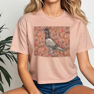 May include: A light pink t-shirt featuring a square graphic of a pigeon wearing a hat. The pigeon is set against a floral patterned background with red, pink, and orange hues. The shirt has short sleeves and a crew neck.