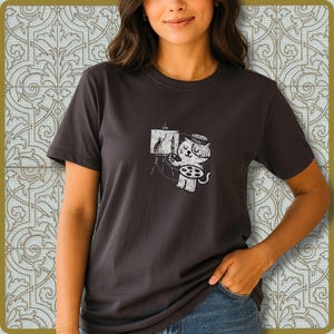 May include: Dark gray t-shirt featuring a whimsical illustration of a cat artist painting a picture of a rabbit on a canvas. The cat wears a beret and holds a paintbrush and palette. The design is in a vintage, distressed style.