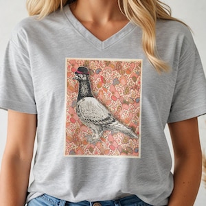 以下が含まれることがあります： 小さな黒い帽子をかぶったハトのグラフィックが特徴の、グレーのVネックTシャツ。ハトは、赤、ピンク、青の花柄の要素を持つパターン化された背景に対して設定されています。シャツはカジュアルなスタイルです。