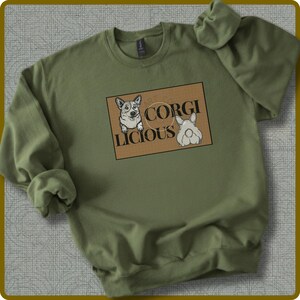 Może przedstawiać: Zielona bluza w kolorze oliwkowym z grafiką z napisem "CORGI LICIOUS". Grafika przedstawia dwa kreskówkowe psy corgi na brązowym prostokątnym tle. Bluza ma długie rękawy i okrągły dekolt.