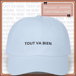 May include: A light blue baseball cap with the phrase "TOUT VA BIEN" embroidered in black. The cap has a curved brim and a rounded crown. The text "Because, ummm, EVERYTHING is FINE!!" is also visible.