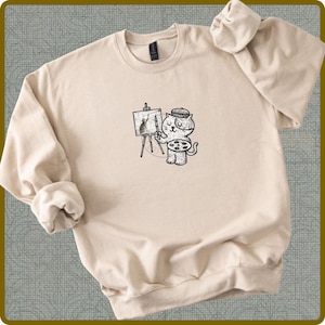May include: A beige sweatshirt featuring a black and white illustration of a cat artist. The cat, wearing a beret, stands before an easel, painting a picture of a rabbit. The sweatshirt has long sleeves and a crew neck.