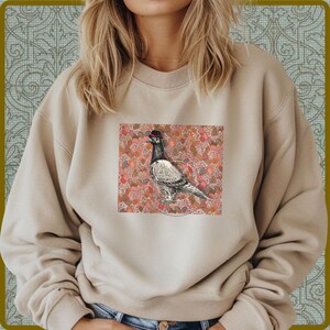 Puede incluir: Sudadera beige con un gráfico de una paloma con bombín. La paloma está sobre un fondo de formas rosas y rojas. La sudadera tiene mangas largas y cuello redondo.
