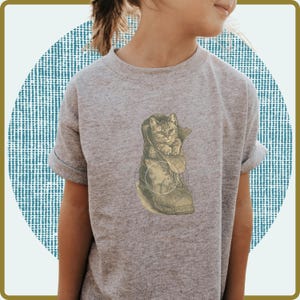 May include: A gray t-shirt featuring a graphic of a kitten inside a boot. The kitten has gray and brown fur. The boot is tan and has a circular design on it. The shirt has short sleeves.