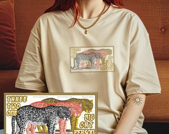 May include: Beige t-shirt featuring a rectangular graphic with three stylized leopards in black, pink, and yellow. The text reads "THREE TWO ONE BIG CAT PERSON ENERGY" in a vintage style. The shirt is a casual, oversized fit.