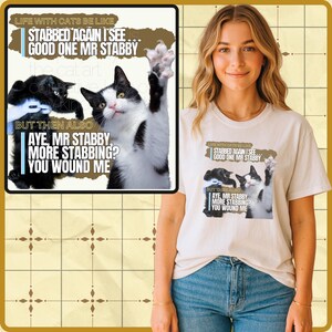 Puede incluir: Una camiseta color crema con un gráfico de dos gatos y el texto "STABBED AGAIN I SEE... GOOD ONE MR STABBY" y "AYE, MR STABBY, MORE STABBING? YOU WOUND ME". La camiseta la lleva una persona.