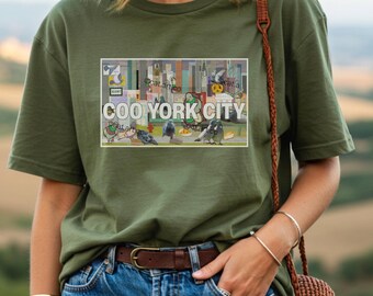 Puede incluir: Camiseta verde oliva con un diseño rectangular que muestra el texto "COO YORK CITY". El diseño representa palomas, edificios y elementos urbanos. La camiseta tiene mangas cortas y cuello redondo.