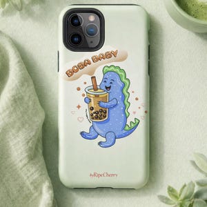 May include: A light green phone case featuring a cartoon blue dinosaur holding a boba drink. The text "BOBA BABY" is above the dinosaur. The case has a camera cutout and the text "byRipeCherry" at the bottom.