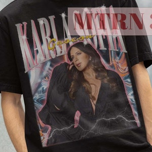 May include: Black t-shirt with a graphic print of a woman with long brown hair. The text "KARLA MORAN" is printed on the shirt. The model is wearing a black bucket hat.