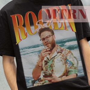 May include: Black t-shirt with a graphic print of a man with a beard and glasses, wearing a floral shirt, sitting on a beach. The text "ROGER" is printed in red, yellow, and orange letters above the image. The text "Seth" is printed in white letters below the image.