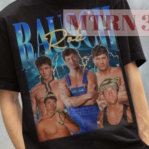 Puede incluir: Camiseta negra con un gráfico de un grupo de hombres y el texto "BASTIEN Rob" en azul y dorado. La persona de la imagen lleva un sombrero de pescador negro.