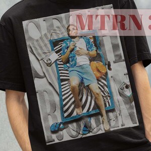 May include: Black t-shirt with a graphic print of a person laying on a beach towel. The graphic includes the text "Surf Rock" and a stylized sun.