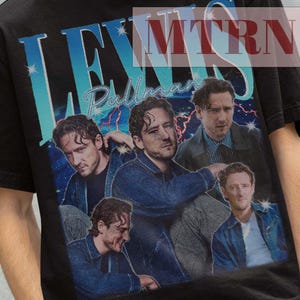 Puede incluir: Camiseta negra con un gráfico de estilo vintage que representa a un grupo de hombres y el texto "LEWIS Pullman" en azul y blanco. La camiseta también tiene el texto "MTRN 38" en rojo. La persona de la imagen lleva un sombrero de pescador negro.