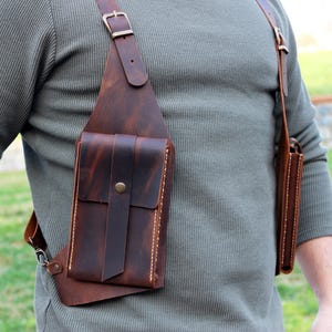 May include: A brown leather crossbody bag with a large pocket on the front and a smaller pocket on the side. The bag is worn over a person's shoulder.