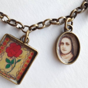 May include: A gold chain necklace with two charms. One charm is a square with a red rose and a yellow background. The other charm is a round oval with a picture of a woman with brown hair and a white collar.
