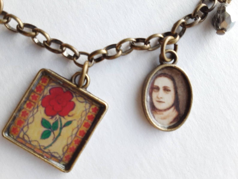 May include: A gold chain necklace with two charms. One charm is a square with a red rose and a yellow background. The other charm is a round oval with a picture of a woman with brown hair and a white collar.