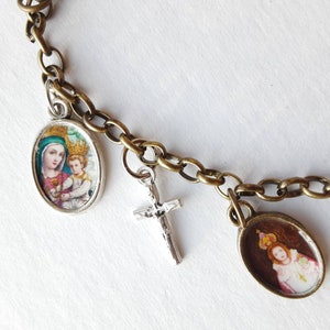 May include: A silver chain bracelet with multiple charms, including a silver cross, a round charm with a picture of the Virgin Mary and baby Jesus, and a round charm with a picture of a young girl wearing a crown.