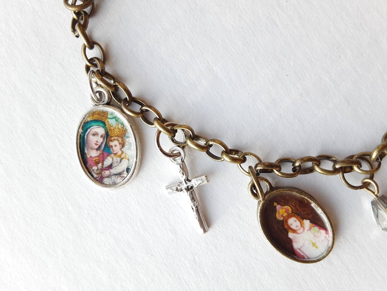 May include: A silver chain bracelet with multiple charms, including a silver cross, a round charm with a picture of the Virgin Mary and baby Jesus, and a round charm with a picture of a young girl wearing a crown.