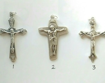 May include: Three silver crucifix pendants with different designs. The first pendant features a figure of Jesus Christ on the cross. The second pendant features a figure of Jesus Christ and Mary on the cross. The third pendant features a figure of Jesus Christ on the cross with a crown of thorns.