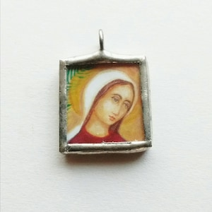 May include: A silver pendant featuring a rectangular glass frame enclosing a painted image of a woman with a white head covering and a red dress. The woman's eyes are closed and she appears to be looking downwards.
