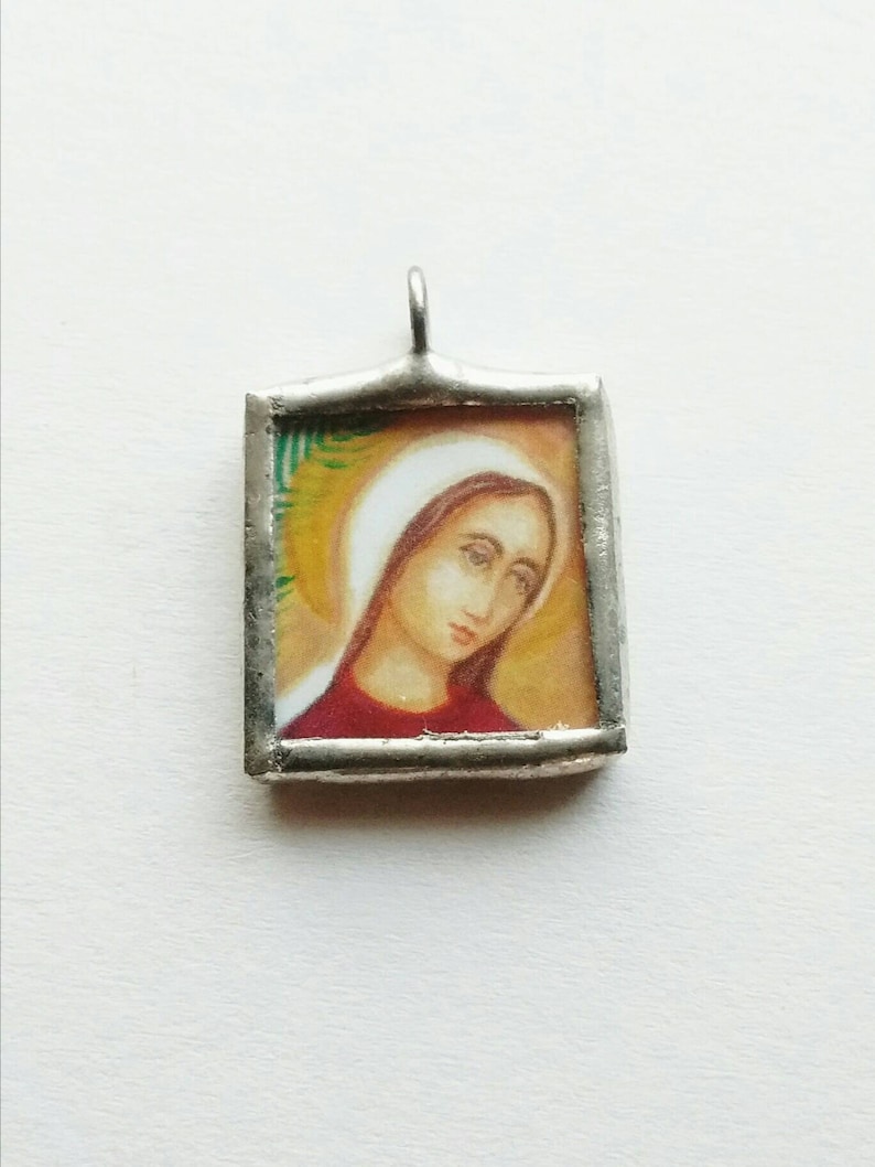 May include: A silver pendant featuring a rectangular glass frame enclosing a painted image of a woman with a white head covering and a red dress. The woman's eyes are closed and she appears to be looking downwards.