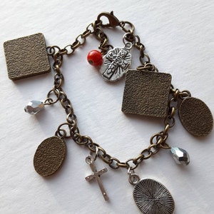 May include: A bronze chain bracelet with multiple charms, including a cross, a red bead, and several square and oval charms. Some charms have a textured surface.