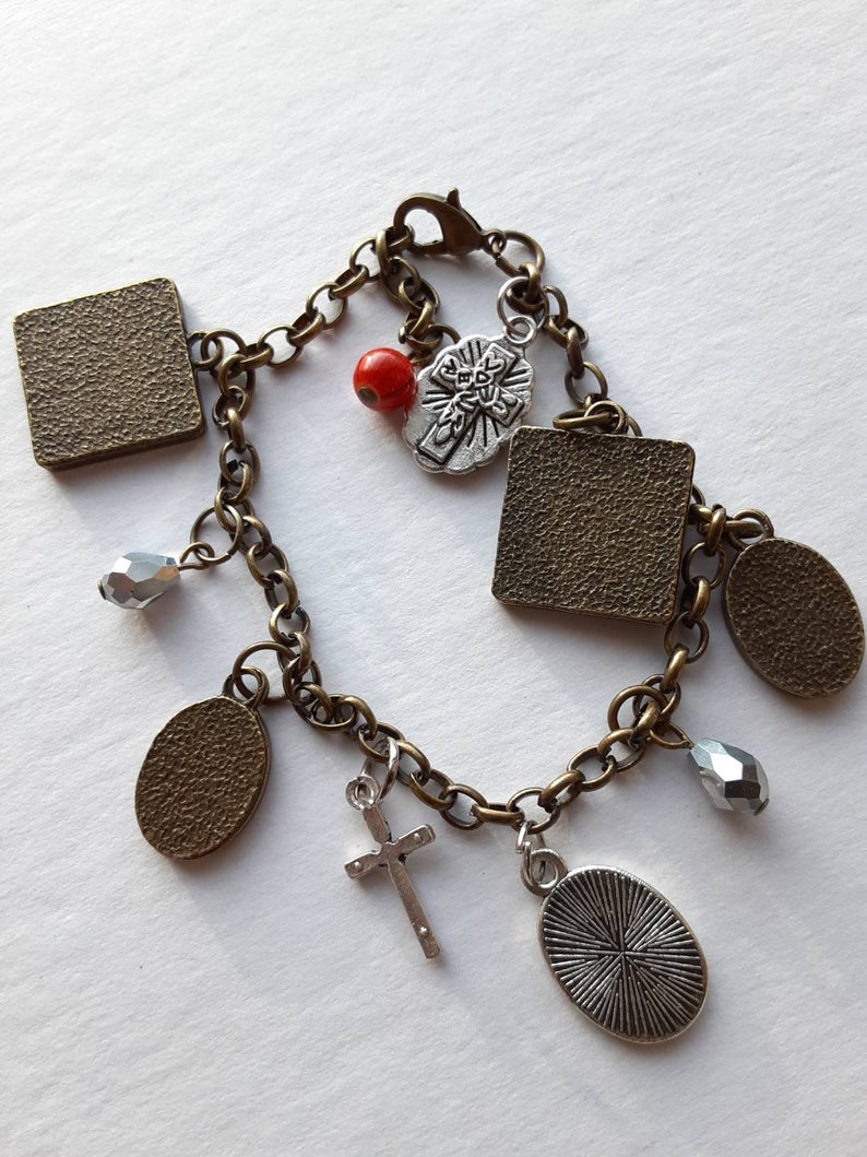 May include: A bronze chain bracelet with multiple charms, including a cross, a red bead, and several square and oval charms. Some charms have a textured surface.