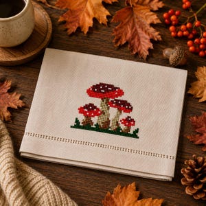 May include: A beige linen napkin featuring a cross-stitch embroidery of four red and white mushrooms with green grass at the base. The napkin has a decorative hemstitch border detail running horizontally across the lower section of the fabric.