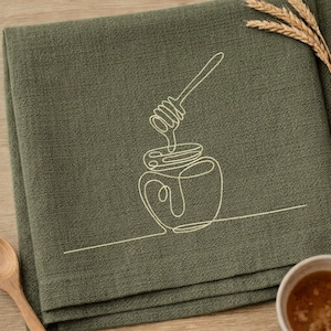 Puede incluir: Servilleta de lino verde oliva doblada con un dibujo lineal blanco de un tarro de miel y un cucharón. Una cuchara de madera y un cuenco pequeño con miel están en el marco. Espigas de trigo en la esquina superior derecha.