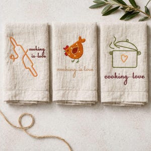 Könnte beinhalten: Drei beige Küchentücher mit gestickten Designs zum Thema Kochen. Eines der Tücher zeigt eine Teigrolle, ein anderes ein Huhn und das letzte einen Topf mit einem Herzen. Jedes Tuch trägt den gestickten Schriftzug "cooking is love".