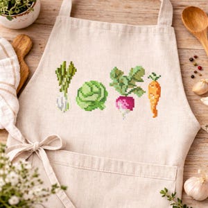 May include: A beige apron with a cross-stitched design of vegetables, including asparagus, cabbage, radish, and a carrot. The apron has a front pocket and a tied bow at the waist. A wooden spoon and small plant are in the background.