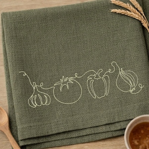 May include: A folded olive green kitchen towel with a white line drawing of vegetables, including a garlic bulb, tomato, bell pepper, and onion. A wooden spoon and a small bowl of soup are in the frame.
