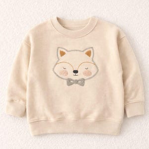 Puede incluir: Sudadera color crema con un bordado de cara de zorro y pajarita gris. El zorro tiene orejas marrones, ojos cerrados y mejillas rosas. La sudadera tiene cuello redondo y mangas largas.