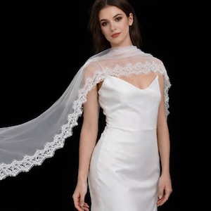 Może przedstawiać: Biała suknia ślubna z dekoltem w kształcie litery V i zwiewnym, przezroczystym welonem. Welon jest obszyty delikatną białą koronką. Suknia wykonana jest z gładkiej, satynowej tkaniny.