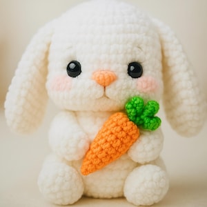 Puede incluir: Un conejito de crochet blanco sosteniendo una zanahoria de crochet naranja con hojas verdes.